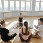 A diverse group of adults meditating in a spacious, sunlit studio with city views.
